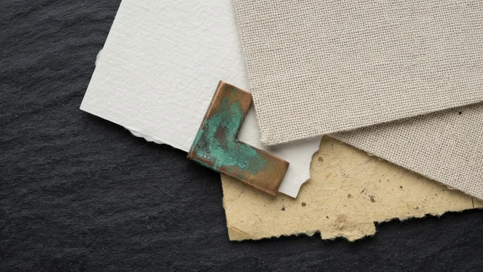 Close view of overlapping paper and canvas offcuts showing cold press grain, fine canvas weave and rough fibrous tooth, pinned by a weathered copper corner piece